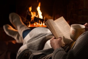 Woman resting with cup of hot drink and book near fireplace