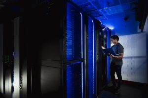 A shot of a young man in a server room with hi-tech supercomputers on display, LED lights are surrounding the equipment he is using.