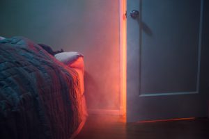 Una representación de un incendio en una casa, en el interior.  El humo y la luz de un incendio se filtran a través de la puerta de una habitación agrietada, se ve a una persona durmiendo en su cama.  Horizontal con espacio de copia.