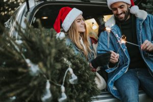 Happy couple is wearing Santa Claus hat and sitting in car trunk while enjoying their beautiful moments of Christmas tree adventure. It is a cold and sunny day outside.