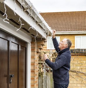 Senior man putting up or taking down outdoor Christmas lights from the gutter of a suburban house